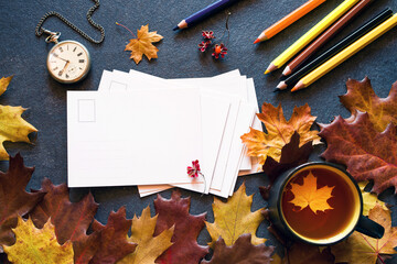 Colorful autumn background with a cup of tea and clean white cards for your text, with bright autumn leaves, pocket watch and colored pencils on a dark background, top view, flat lay.