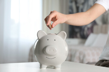 Woman putting money into piggy bank at white table indoors, closeup