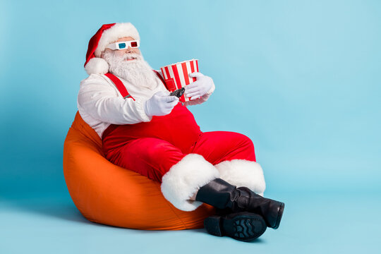 Full Length Photo Of Retired Old Man Grey Beard Hold Remote Controll Bucket Watch Sit Orange Beanbag Wear Red Santa X-mas Costume Suspender 3d Glasses Cap Boot Isolated Blue Color Background