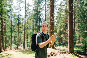 Young tourist with a serious face and in casual clothes stands in woods with a smartphone in his hands, looks away and uses a map on his phone.Hipster stands in the woods on a hike,uses a smartphone