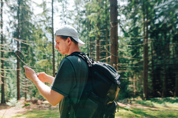 Young hiker man in a cap and casual clothes carries a backpack, stands in a pristine forest and watches the route on maps in a smartphone.Tourist uses the Internet on a smartphone for a walk in woods