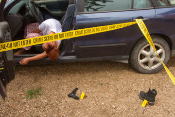 forensic investigations in progress with scientific evidence signs and yellow police tape at a fake corpse of a woman behind the wheel of her car with her head and hands out the car door and slumped