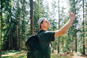 Naklejka na ściany i meble Young man stands in a beautiful mountain forest and catches a mobile network on a smartphone with serious face.Tourist looking for a cellular connection on a hike, raised his hand with a smartphone up