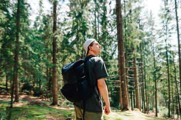 Young male tourist with a backpack and a cap stands in a sunny forest and looks away with a serious face. Guy walks a day in the woods and looks at the beautiful views. Recreation