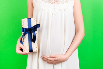 Cropped image of pregnant woman in white dress holding a gift box and touching her belly at green background. Expecting a baby boy. Copy space