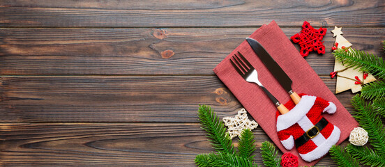 Top view of fork and knife put in santa clothes on napkin with christmas decorations and new year tree on wooden background. Banner holiday and festive concept with copy space