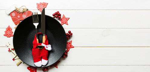 Holiday composition of plate and flatware decorated with Santa clothes on wooden background. Banner Top view of Christmas decorations with empty space for your design. Festive time concept