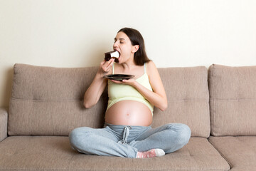 Hungry pregnant woman is eating a piece of tasty cake relaxing on the sofa at home. Sweet cravings during pregnancy