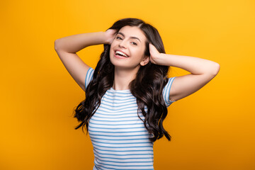 brunette woman toughing curly long hair and smiling isolated on yellow