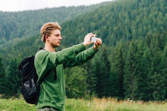 Young male hiker stands in the mountains on a background of hills with coniferous forest and makes photo landscape on a smartphone. Hipster tourist with a backpack takes photo on a walk in mountains.
