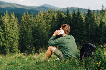 Naklejka na ściany i meble Man talking on the phone while sitting in a clearing among the trees, looking at the summer landscape. Young tourist calling by phone sitting on a meadow in the mountains, looking aside at copy space.