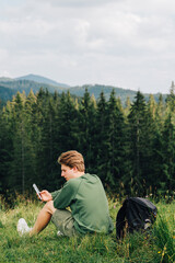 Naklejka na ściany i meble Man sitting on the background of a mountain landscape checking his smartphone. Hiker uses internet on smartphone in mountains during vacation, sitting on meadow against mountains and mountain forest
