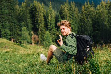Young man smiling sits on a mountain meadow among the forest. Joyful young male hiker resting on the grass in the mountains and showing OK gesture with his fingers.
