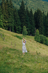 Photo of a beautiful woman in a dress posing for the camera on a green meadow in the mountains with arms raised. Attractive girl stands in the mountains and enjoys nature