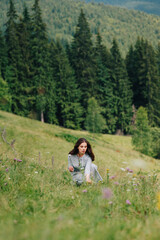 Pretty girl in a blue dress collects flowers on a meadow in the mountains. Attractive lady gathers grass in the mountains. Vertical photo