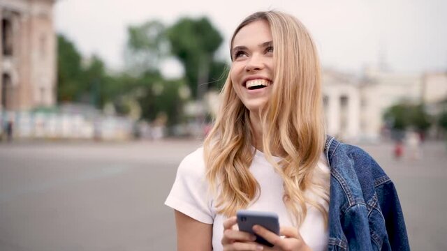Young pretty cheerful girl looking at the phone and walking down the street. Front view, dolly shot. Happy blonde female student on campus.