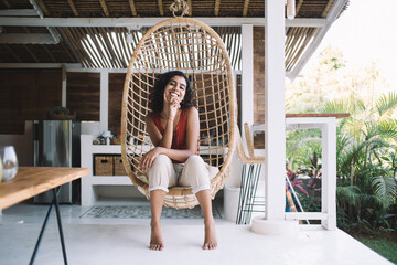 Portrait of joyful woman in casual clothing smiling at camera during rest swinging in rattan hanging chair, pretty Middle Eastern female relaxing in wicker furniture during weekend at porch - Powered by Adobe