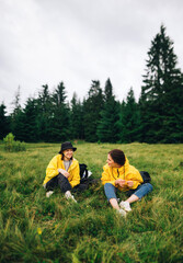 Naklejka na ściany i meble Vertical photo of two happy tourists resting sitting on a meadow in the mountains. Pass on the grass in the mountains in two positive women. Mountain hike