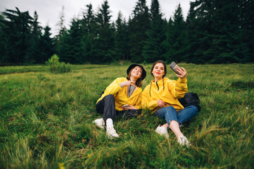 Naklejka na ściany i meble Two cheerful girls tourists are sitting on a mountain meadow on a hike and taking selfies on a smartphone camera.Attractive women in yellow raincoats take selfies resting on the grass in the mountains