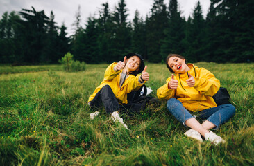 Two happy women in a mountain hike sitting on the grass on a pipe against the backdrop of the forest, looking at the camera and smiling, pointing thumbs up, wearing yellow raincoats.