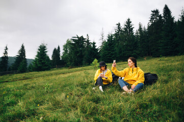 Two girls tourists in yellow raincoats on a hike sitting on a meadow in the mountains and using smartphones. Female hikers with smartphones in their hands resting on the pass sitting on a meadow.