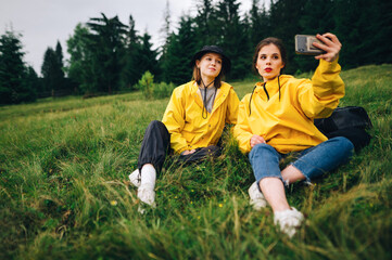 Naklejka na ściany i meble Beautiful girl in a yellow raincoat taking a selfie on a mountain hike sitting on a meadow on a background of forest. Tourists take a selfie at a halt during the hike
