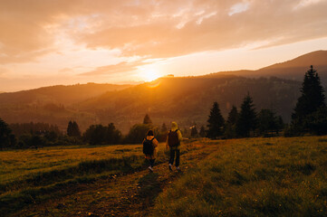 Two tourists in raincoats descend from the mountain in rainy weather against the backdrop of an unreal sunset. Hikers walk down the meadow on a background of sunset in the rain and mountain landscape