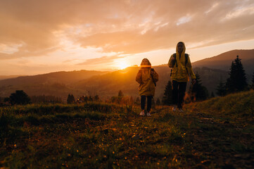 Two women in raincoats on a hike climb a mountain in rainy weather with backpacks on their backs. Girls hikers go to the mountains in the evening at sunset in raincoats during the rain