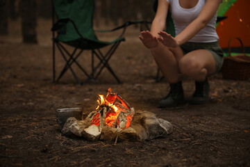 Woman warming hands near burning firewood in forest, closeup