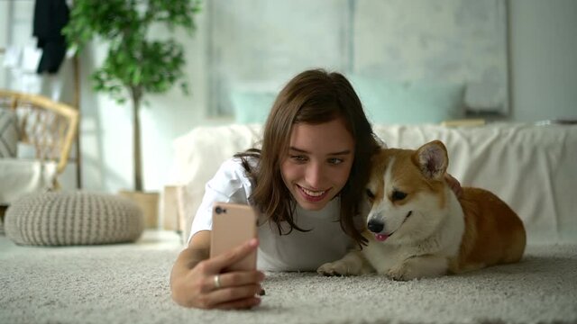Young beautiful woman taking selfie photo with dog and lying on floor in home interior avki. Front view of American girl makes picture with smile and hugs cute pet while holding smartphone in hand