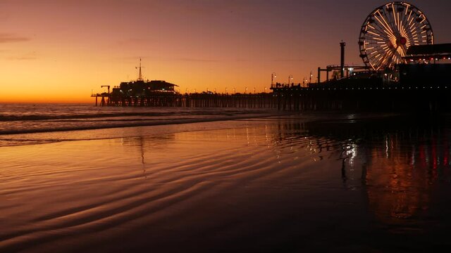 Twilight waves against classic illuminated ferris wheel, amusement park on pier in Santa Monica pacific ocean beach resort. Summertime iconic symbol of California glowing in dusk, Los Angeles, CA USA