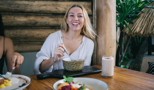 Portrait of cheerful blonde young woman looking at camera enjoying tasty food on breakfast keeping vegan diet, cropped image of hipster girl eating meal during lunch satisfied with dishes and drinks