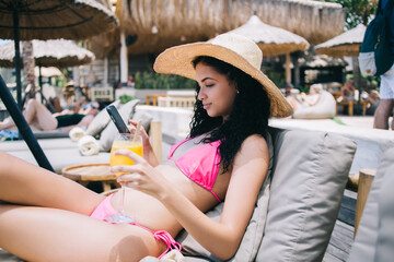 Portrait of serious caucasian female in stylish swimsuit and hat recreating on villa resort with drink using mobile phone for chatting, woman tourist looking at camera sending multimedia in blog