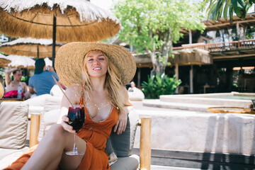 Positive caucasian blonde in dress and trendy hat lying on sunbed during free time vacations on resort, attractive cheerful woman looking at camera holding glass with cold beverage cocktail relaxing