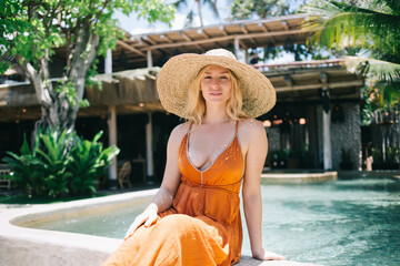 Portrait of attractive blonde caucasian woman in trendy hat and dress recreating near poolside on...