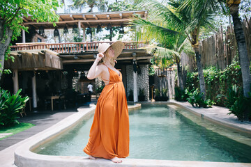 Beautiful blonde female in trendy hat and dress with belly expecting baby spending free time on...