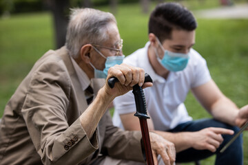 Two generations, a grandson and a grandfather, sit on a bench using a tablet and talking under masks