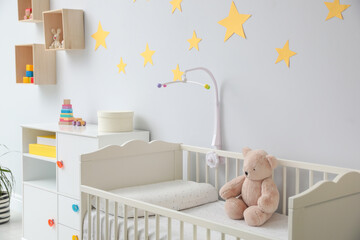 Crib with toy bear and mobile in stylish baby room interior