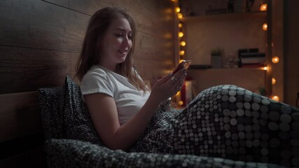 Happy smiling young woman texting on smartphone in bed at home bedroom at night. technology, Internet, communication and people concept .