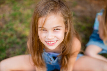 a little girl with long hair in a denim jumpsuit sits on the grass and looks up. happy carefree childhood.