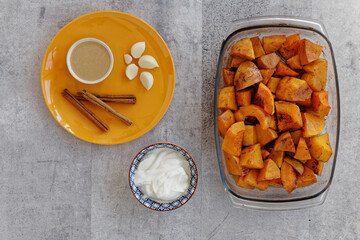 Baked in glass dish butternut squash with cinnamon and other ingredients (tahini paste, garlic, yoghurt) to prepare mashed butternut squash spread. Orange color plate and gray background, flat lay.