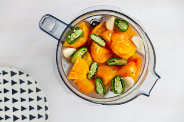 Blender cup with yellow tomatoes, green chili and garlic to make simple and fast tomato sauce. Top view,  triangle patterned plate next to object.