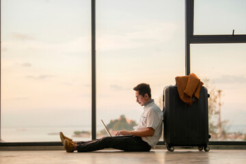 Work remote. Handsome young man working and typing on laptop while sitting on the floor leaning on suitcase near big window at airport and Background beautiful sunset...