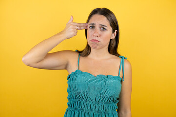 Young beautiful woman over isolated yellow background hooting and killing oneself pointing hand and fingers to head like gun, suicide gesture.