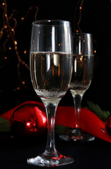 Two glasses of champagne on the festive table on a dark background. next to Christmas decor red balls, ribbon, bright garland in the background. Christmas composition. Selective focus