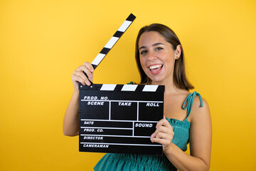 Young beautiful woman over isolated yellow background holding clapperboard very happy having fun
