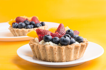 2 tartaleas tropicales de frutas: melocotón, fresas y arándanos sobre fondo de color amarillo.