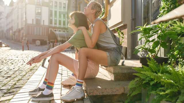 Young lesbian couple, two girls in casual wear and rainbow colored socks smiling, sitting on the steps while having a walk around the city. LGBT, relationship concept