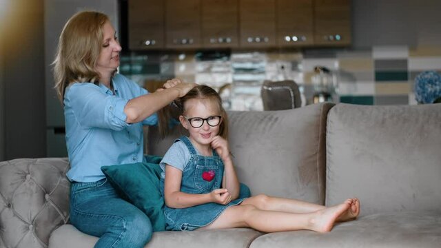 mother preparing daughter for school by doing ponytail hairstyle Rbbro. care for child. mom sit on couch and make girl hair. kid smiles and talk. morning routine. concept love, help