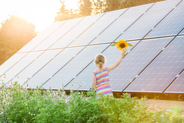 Young 6 year old blonde girl child standing in front of small solar panel farm in countryside....